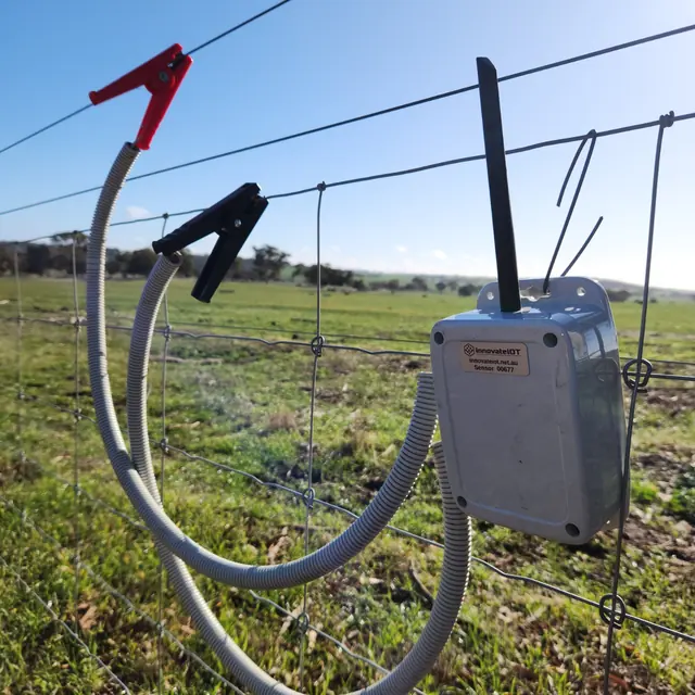 The electric fence monitor hanging on a wire fence, the red clip on the live wire, with open paddock behind