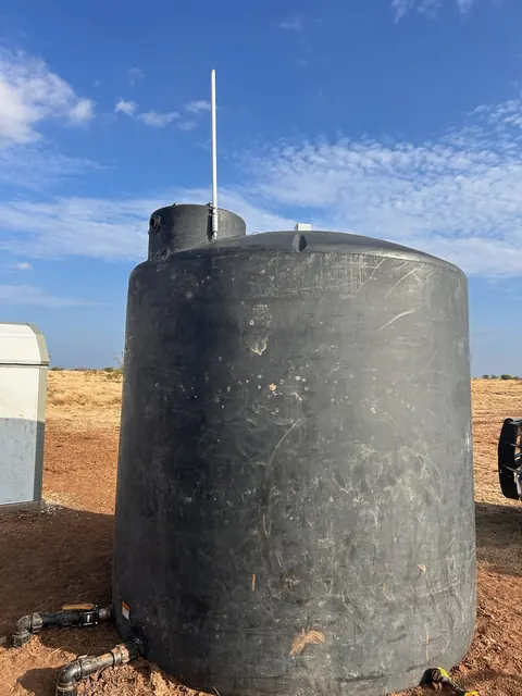 A large black poly water tank on dry rangeland with the sensor on top, a white external antenna above the fill collar, and an equipment cabinet beside it
