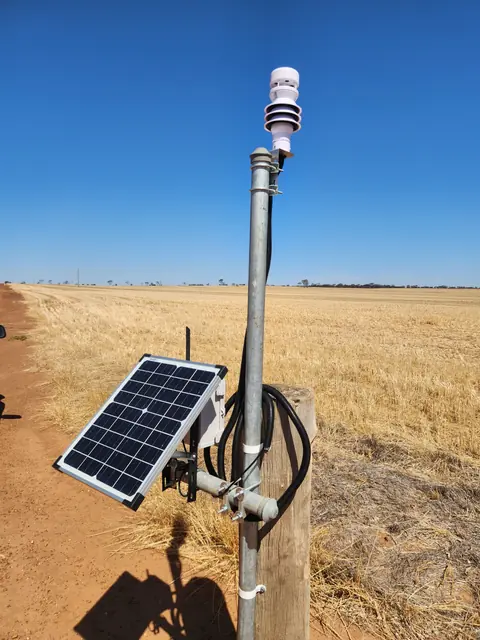 A weather station on a steel post in a harvested paddock: the sensor head on top, solar panel and communications box lower on the post