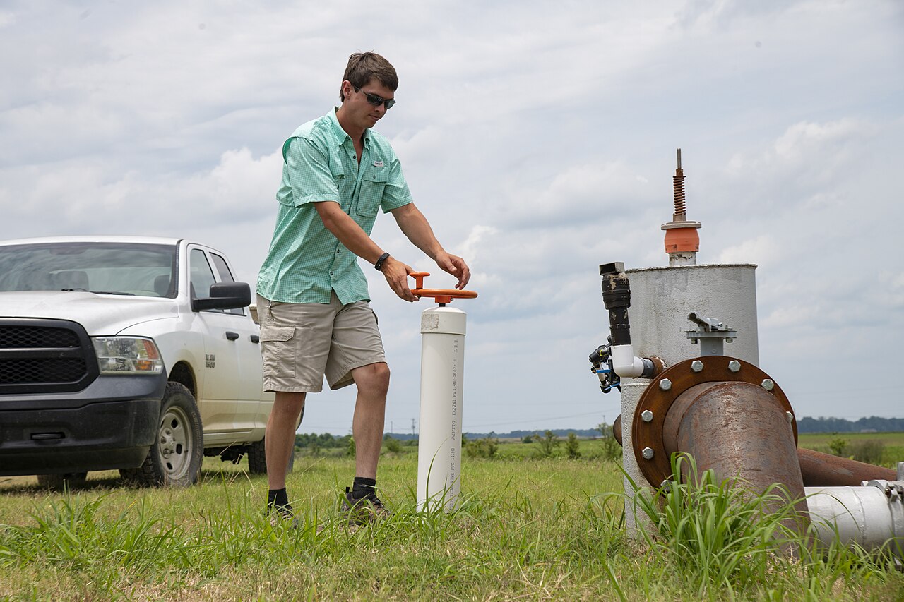 A farmer operates a water-control riser beside an irrigation pump on a farm