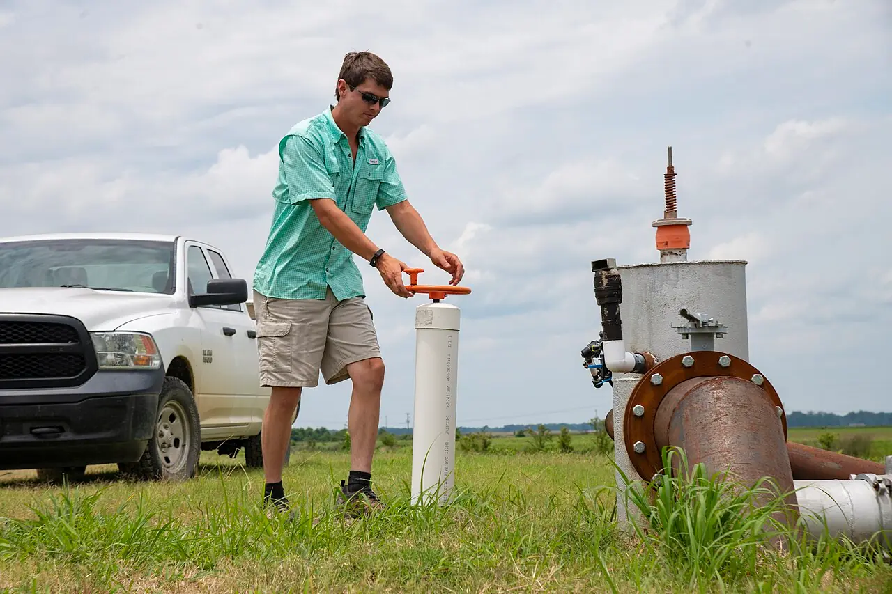A farmer operates a water-control riser beside an irrigation pump on a farm