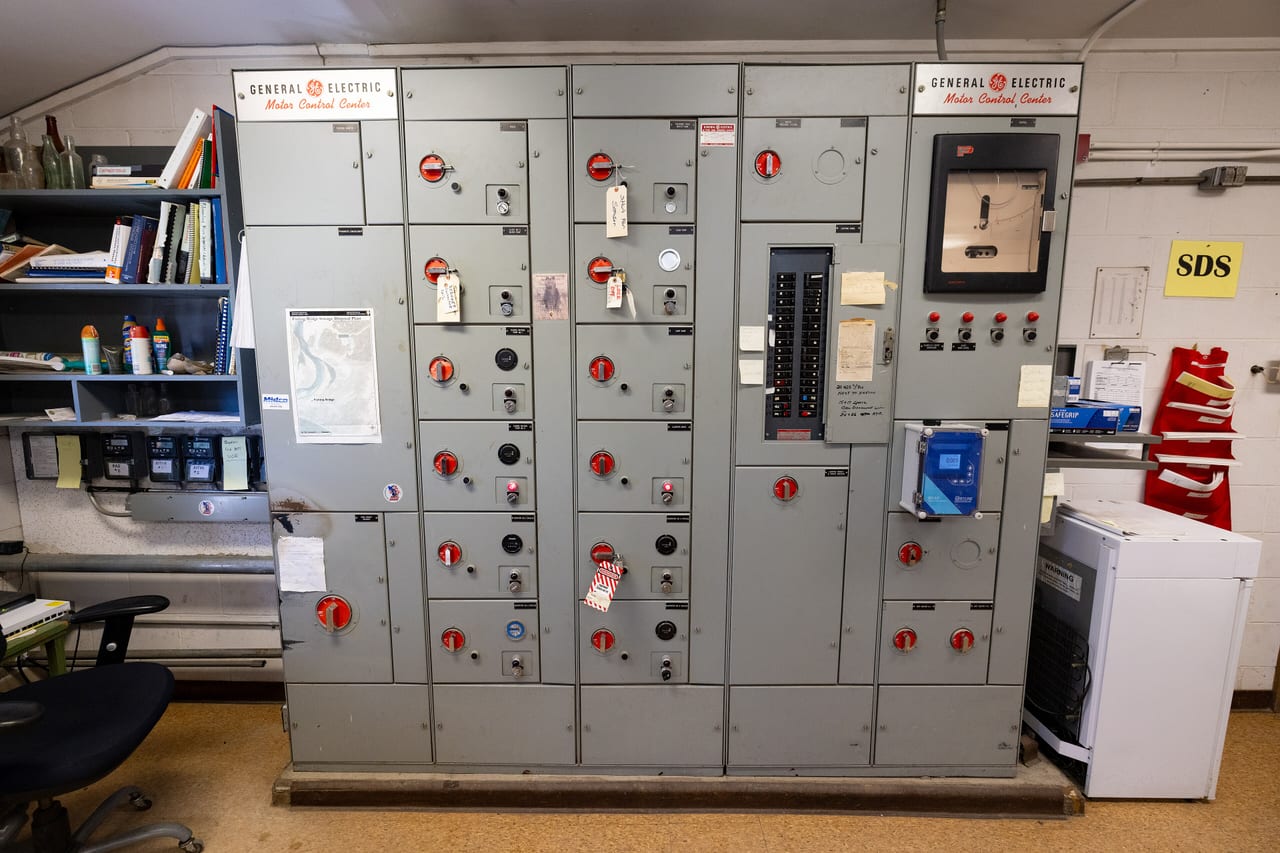 A grey motor control center cabinet lining a plant room wall, with rows of red disconnect handles, meters, and handwritten labels