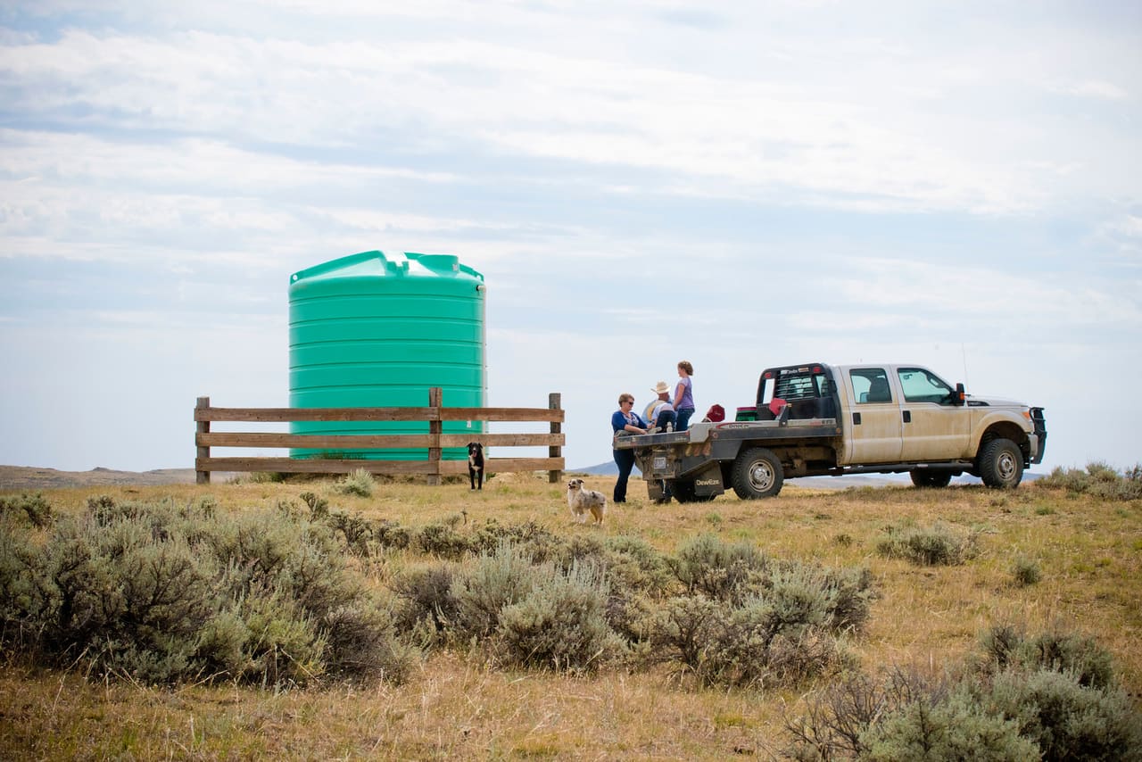 Green poly water storage tank behind a rail fence on sagebrush rangeland, ranch family at a flatbed truck nearby