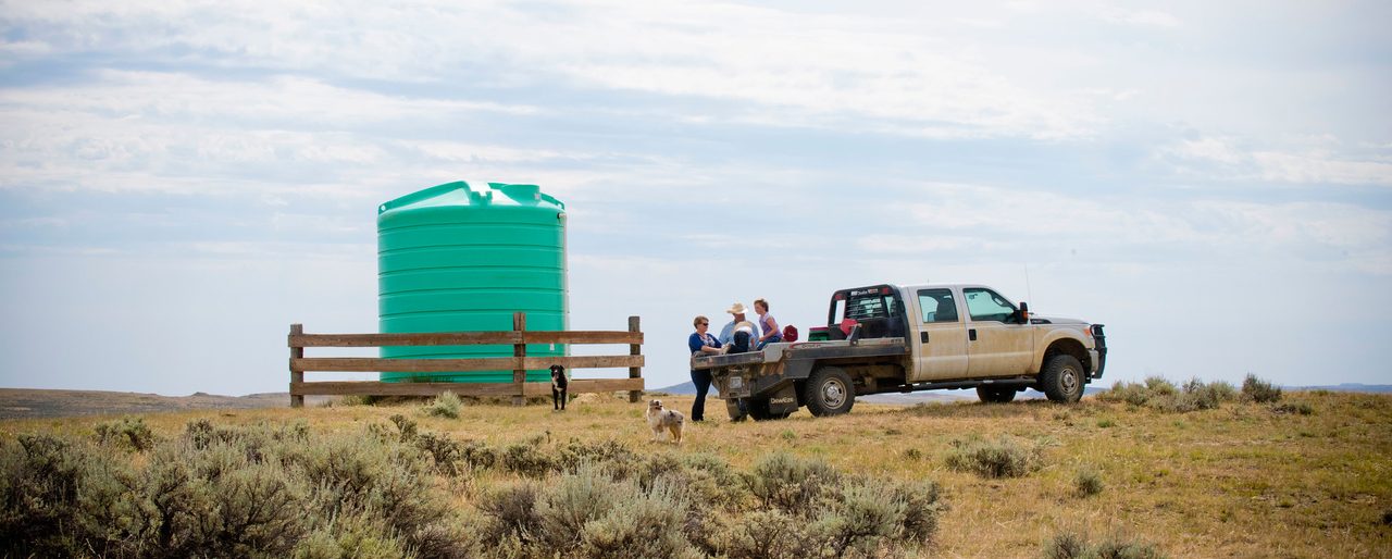 A large poly water storage tank behind a windbreak fence on open rangeland, with a ranch family beside their flatbed truck