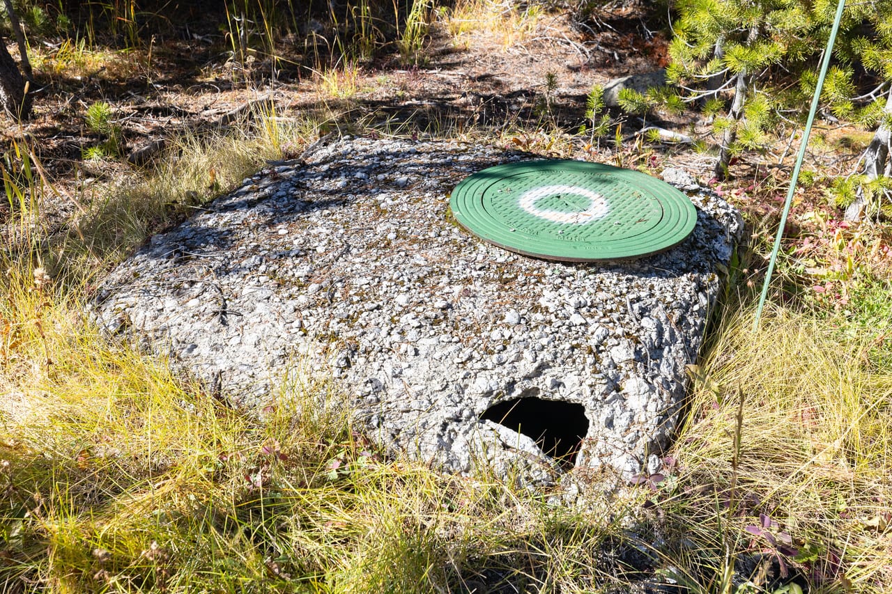 A concrete sewer access point in rough ground with its surround eroded through, leaving an open gap beside the lid