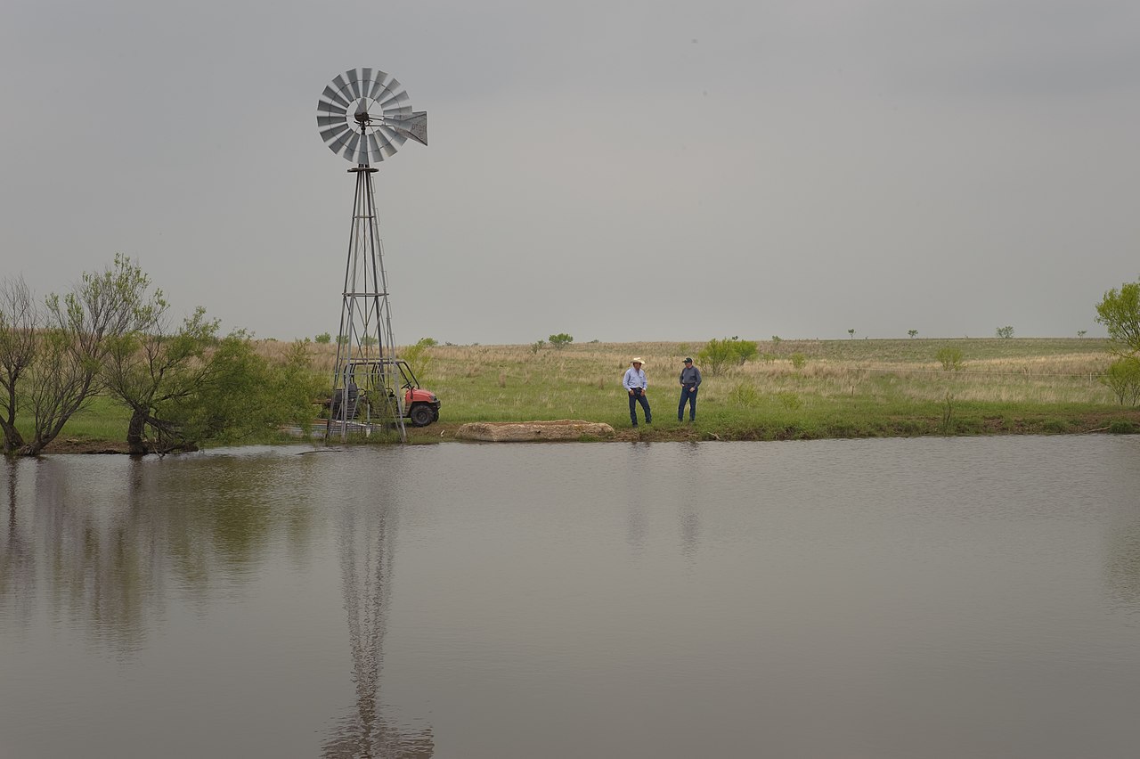 A windmill and stock pond on open rangeland, with two ranchers standing on the bank