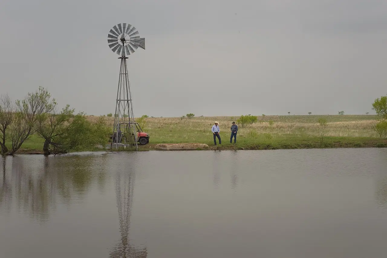A windmill and stock pond on open rangeland, with two ranchers standing on the bank