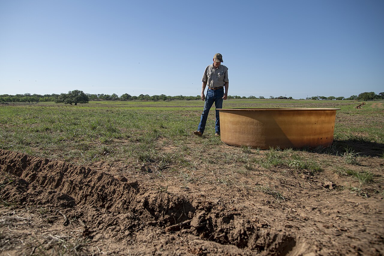 A rancher stands beside a large galvanized steel stock tank on a ranch pasture