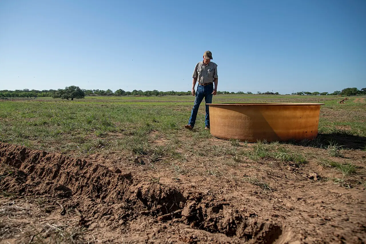 A rancher stands beside a large galvanized steel stock tank on a ranch pasture