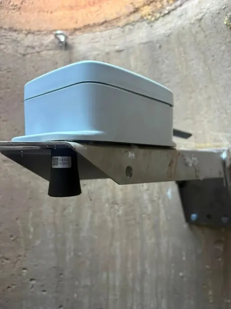 The grey monitor enclosure on a steel bracket bolted to the inside wall of a manhole, with the ultrasonic sensor pointing down